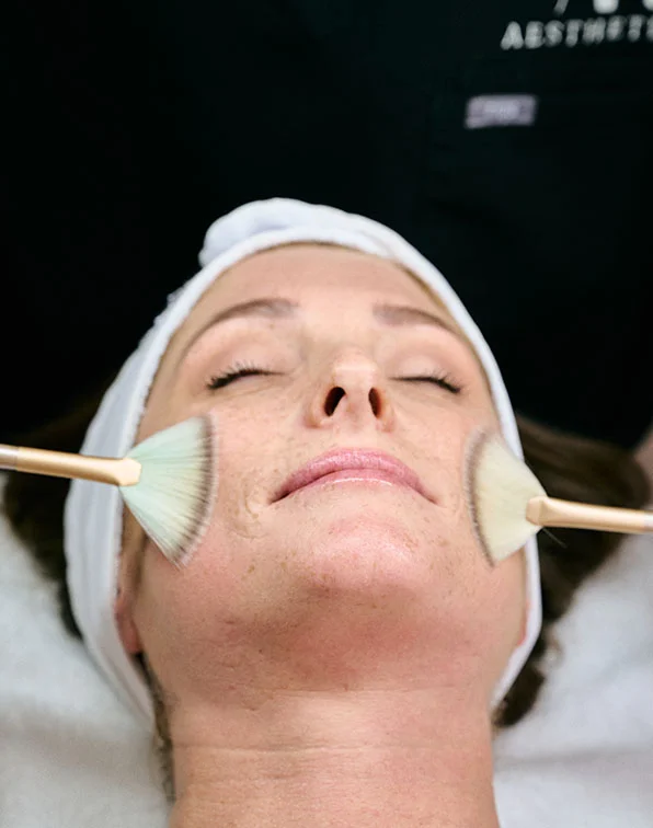 A woman lies with her eyes closed, receiving a facial treatment. A light beige/tan substance is being applied to her cheeks using large, fan-shaped brushes. The brushes are light beige/gold in color. The woman is wearing a white head wrap. Her skin appears to be light-toned and has some visible fine lines and freckles. The background is dark, possibly a dark gray or black, with a blurred appearance, suggesting a spa or salon setting. A portion of a person's arm or shoulder in a dark garment is slightly visible in the background. - Acne Scarring in Tampa, Melbourne and St. Petersburg, FL
