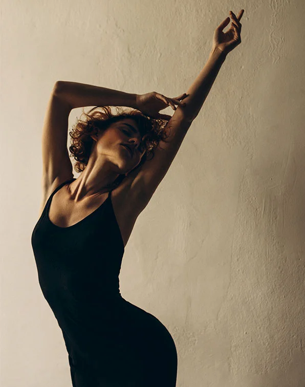 A woman with curly brown hair poses fluidly against a textured light beige wall. Her arms are outstretched at different heights, and her expression is relaxed and contemplative. She wears a form-fitting black sleeveless dress that highlights her silhouette. Soft lighting emphasizes her curves, adding warmth to the scene. - Armsculpt in Tampa, Melbourne and St. Petersburg, FL