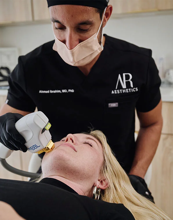 A light-skinned woman lies on a treatment table with her eyes closed while Dr. Ibraham, wearing a black “AR AESTHETICS” scrub top and a light beige face mask, performs a facial treatment. He uses a white and gray device, likely a laser, along her jawline. The woman, dressed in black, appears relaxed as he focuses on the procedure. The background is light beige. - BBL IPL in Tampa, Melbourne and St. Petersburg, FL
