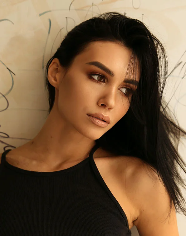 Close-up of a woman with dark, tousled hair cascading over her back and shoulders. She has smooth, tan skin and wears a black halter top. Her expression is neutral, facing slightly left, with warm-toned makeup in taupe and nude shades. The background is a light beige wall with faint scribbled patterns, softly lit. - Botox Dysport Xeomin in Tampa, Melbourne and St. Petersburg, FL
