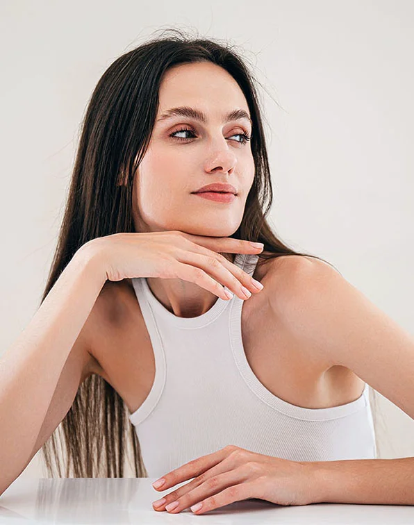 A light-skinned woman with long dark brown hair sits angled slightly right, gazing off to the side. She rests her hands gently on her chin and a table, wearing a plain white ribbed tank top. The soft beige background and even lighting highlight her smooth skin and natural makeup. Her nails are light beige or pinkish, creating a relaxed, thoughtful portrait. - Deep Laser Skin Resurfacing in Tampa, Melbourne and St. Petersburg, FL