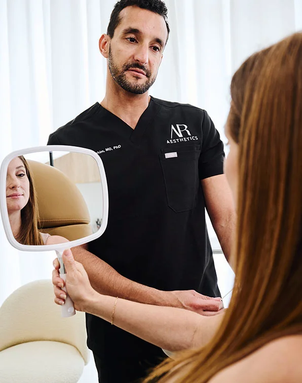 A medium shot shows a doctor-patient consultation. A light-skinned man in a black scrub top with "AR Aesthetics" embroidered holds a white handheld mirror for a woman with shoulder-length light brown hair. She holds the mirror and looks into it, seeing her reflection. The scene takes place in a professional medical office with light walls, beige furniture, and sheer white curtains in the blurred background. - Dimpleplasty in Tampa, Melbourne and St. Petersburg, FL
