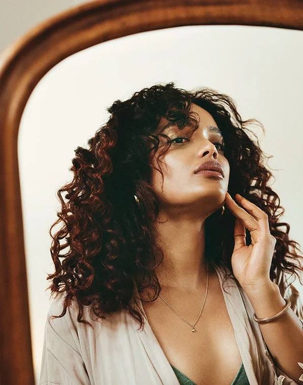 A woman with curly dark brown hair gazes at herself in a mirror, wearing a light beige robe and a gold-toned necklace. A hint of a greenish-grey undergarment is visible beneath. She touches her neck and cheek thoughtfully. The mirror has a light brown frame, and the background is a soft cream color with gentle, diffused lighting. - PRP PDGF Injections in Tampa, Melbourne and St. Petersburg, FL