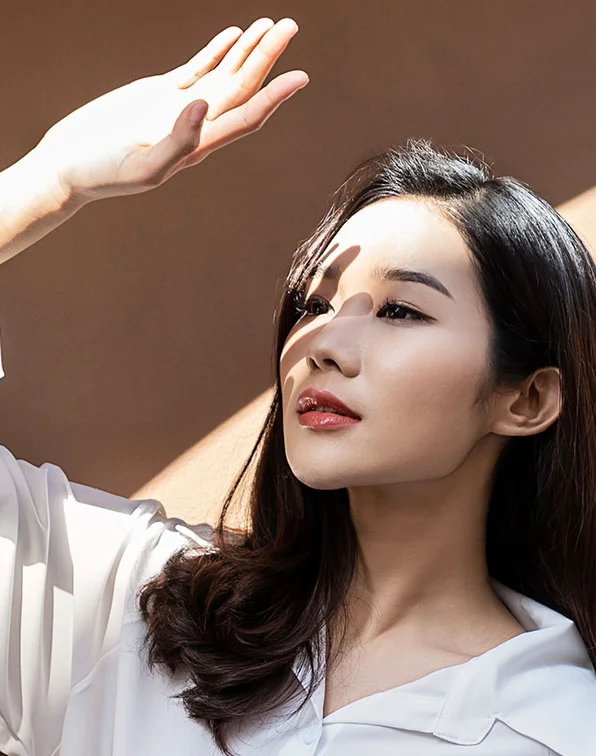 A close-up of a woman with long, dark brown hair wearing a white satin button-down shirt. Her arm is raised as if shielding her face from sunlight, casting a soft shadow across her features. Her expression is neutral, eyes looking toward the light, with smooth light-toned skin and pink-nude lips. The muted beige background keeps focus on her face and gesture. - Revivelift in Tampa, Melbourne and St. Petersburg, FL