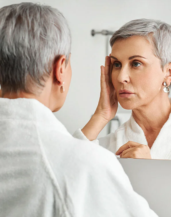 A woman with short, gray hair is looking at her reflection in a mirror. She is wearing a white robe. She is touching her face, likely examining her reflection. The woman in the mirror appears to have some visible signs of aging, such as wrinkles. The setting appears to be a bathroom or similar space. The focus is primarily on the woman and her reflection, with a soft out-of-focus background. - Sclerotherapy in Tampa, Melbourne and St. Petersburg, FL