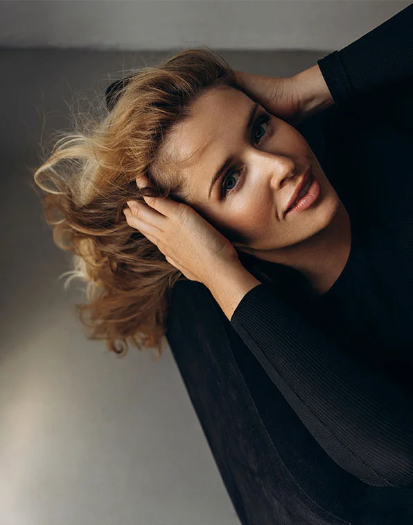 A woman with shoulder-length, tousled light brown hair is seen from a high angle, head tilted back and hands holding her hair. She wears a fitted black long-sleeve top against a muted light-gray background. Soft lighting highlights her pensive expression, with eyes looking upward and lips a natural pink. - Surgical Facial Scar Revision in Tampa, Melbourne and St. Petersburg, FL