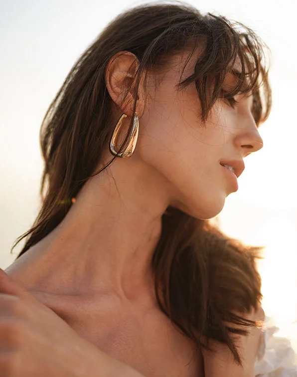 A close-up profile of a woman with long, dark brown hair and a light tan complexion. She wears large gold-toned earrings and a light-colored top. Her expression is serene as she gazes off to the side. Soft sunlight highlights her skin and hair, with a pale, blurred outdoor background. - TMJ Botox in Tampa, Melbourne and St. Petersburg, FL